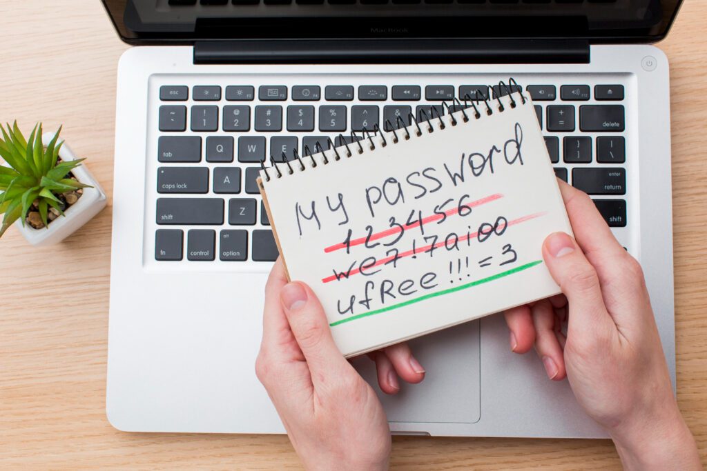 A person holding a notebook with different crossed-out passwords on top of a laptop